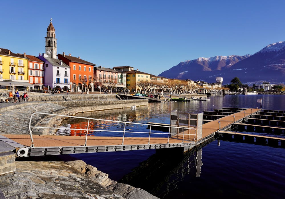 Ascona: Malerisk promenade ved bredden med farverige bygninger og bjerge i baggrunden.