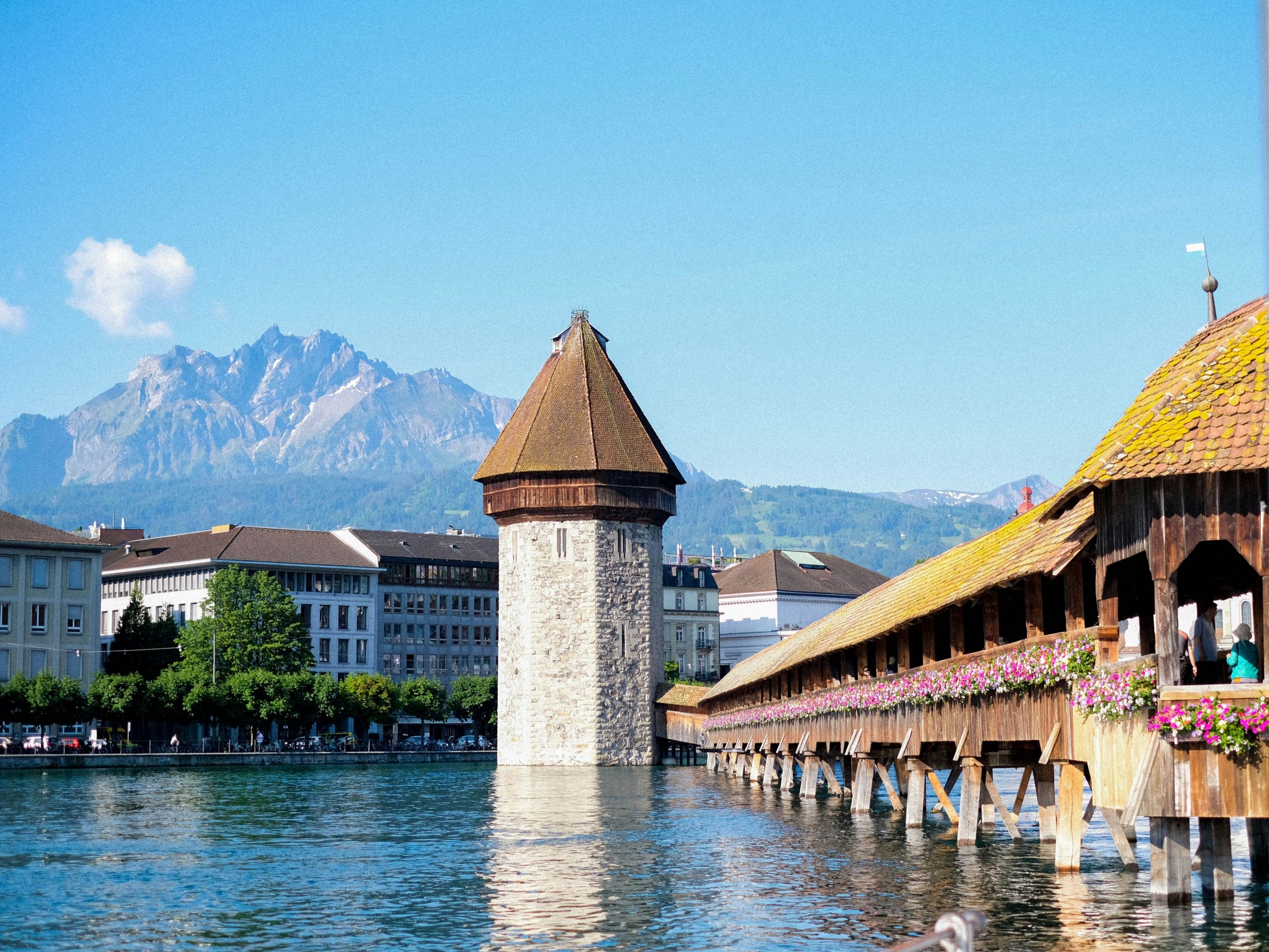 Lucerna: Immagine della Torre dell'Acqua e del Ponte della Cappella sotto il sole di Lucerna con le montagne sullo sfondo.