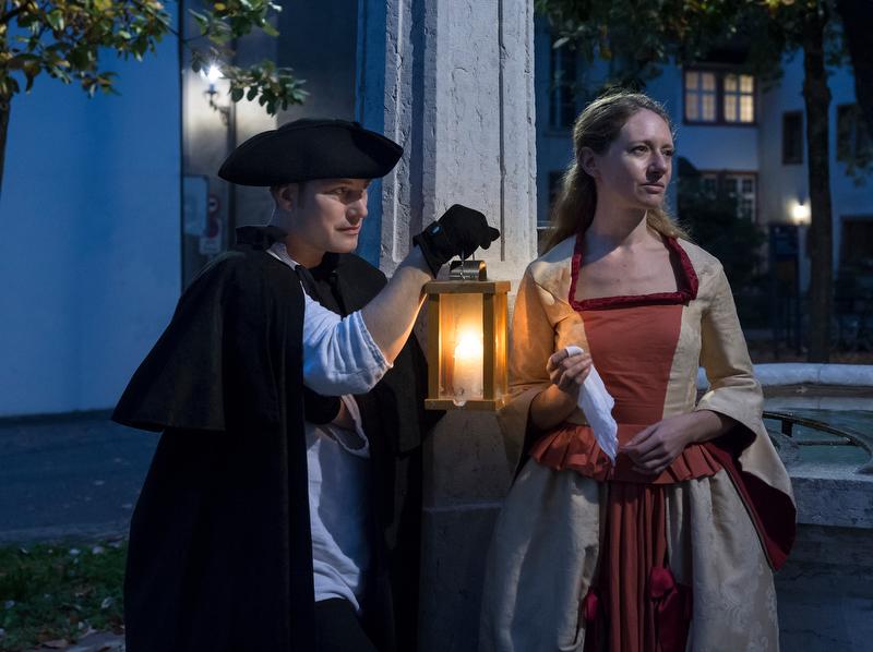 Lawatan malam di Basel dengan tokoh bersejarah di hadapan lampu jalan.