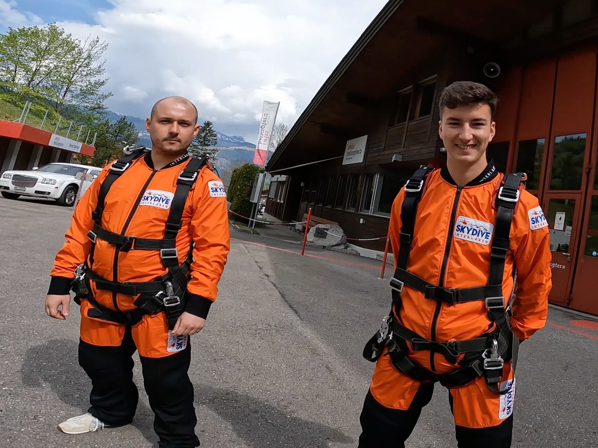 Skydive Interlaken: สองคนในชุดกระโดดสีส้มที่ฐานเฮลิคอปเตอร์ ล้อมรอบด้วยภูเขาและท้องฟ้าที่แจ่มใส.
