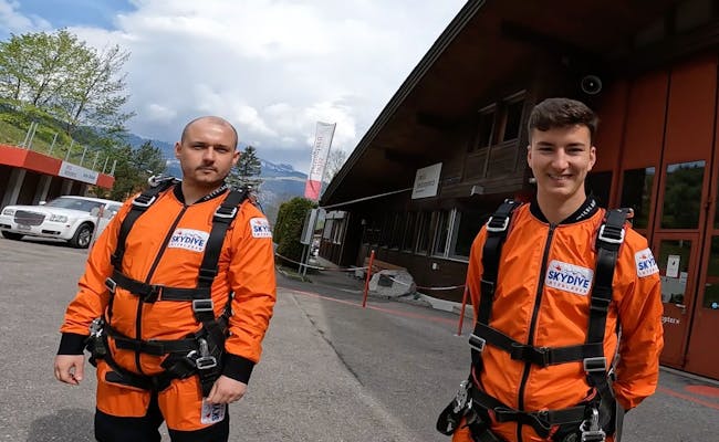 Skydive Interlaken: Zwei Personen in orangefarbenen Sprunganzügen an der Helikopterbasis, umgeben von Bergen und einem klaren Himmel.