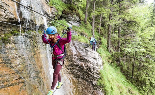 Jalur Panjat Tebing Mürren dengan orang-orang di dinding batu, dikelilingi oleh pohon dan air terjun