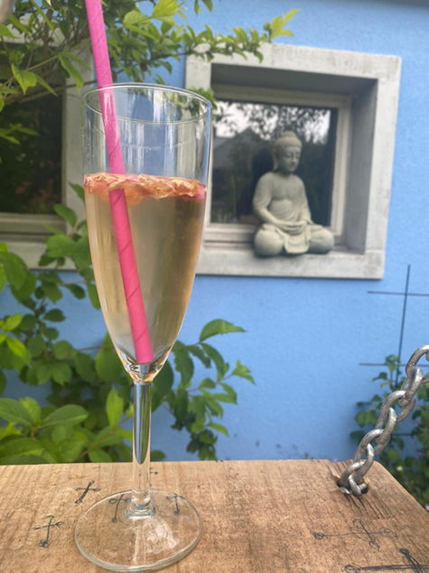 Glas mit Getränk und rosa Strohhalm im Garten vor Buddha-Statue.