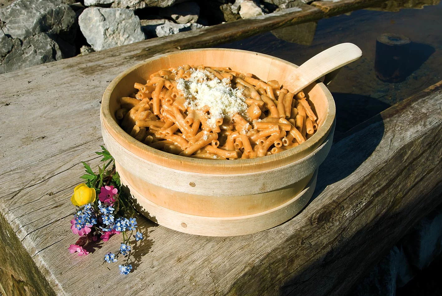 Älpler Magronen v dubové misce se sýrem na dřevěných prknech, obklopené květinami