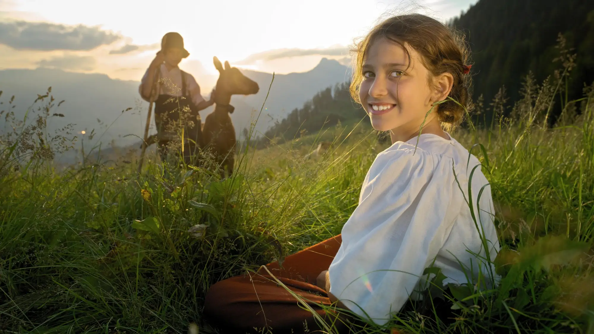 ハイジは笑顔で草の上に座っていて、背景には動物を持った人が見えます。