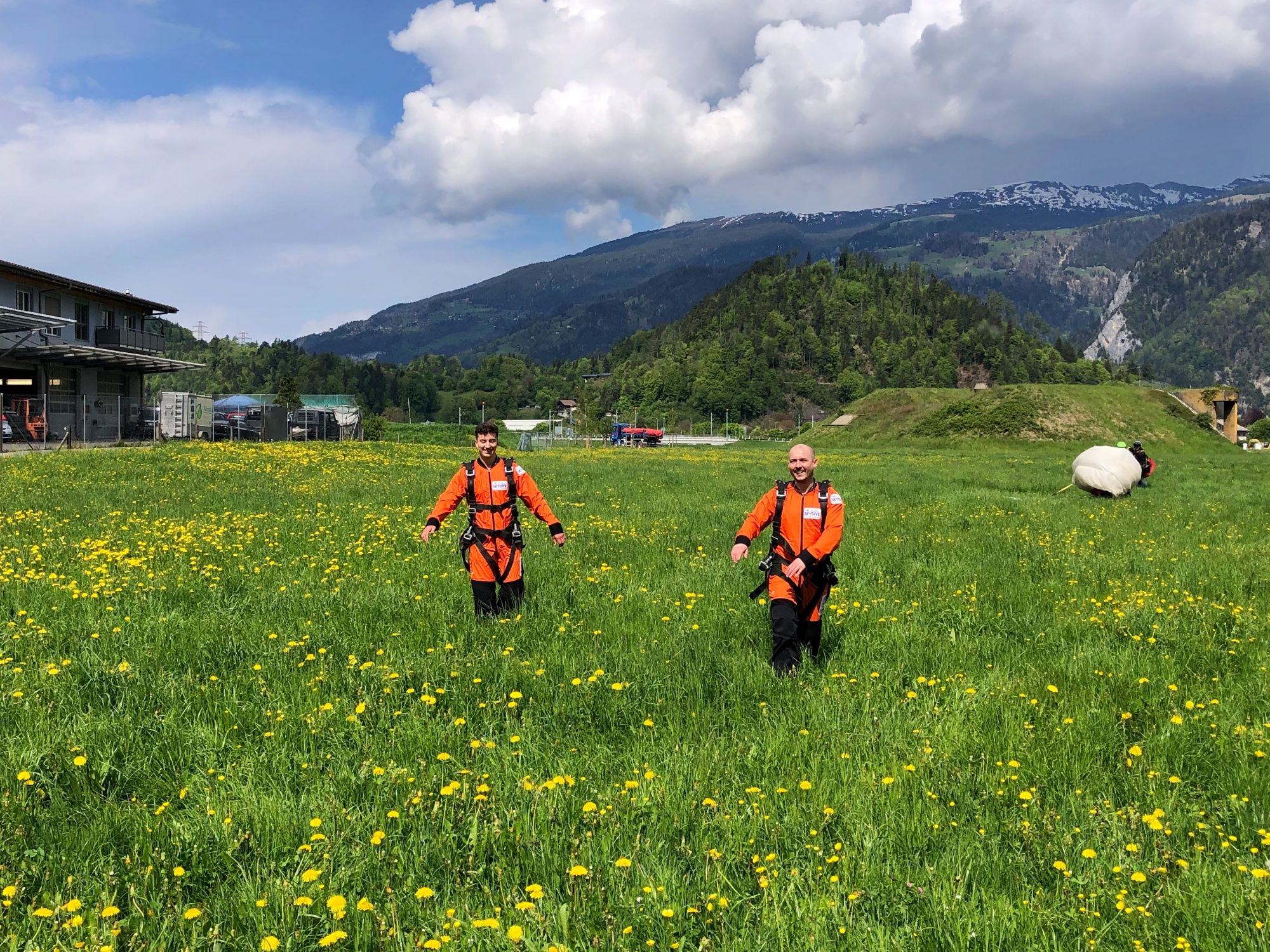 นักบินสกายไดฟ์สองคนในชุดสีส้มกำลังเดินอยู่บนทุ่งหญ้าที่เบ่งบานในเมืองอินเทอร์ลาเคน