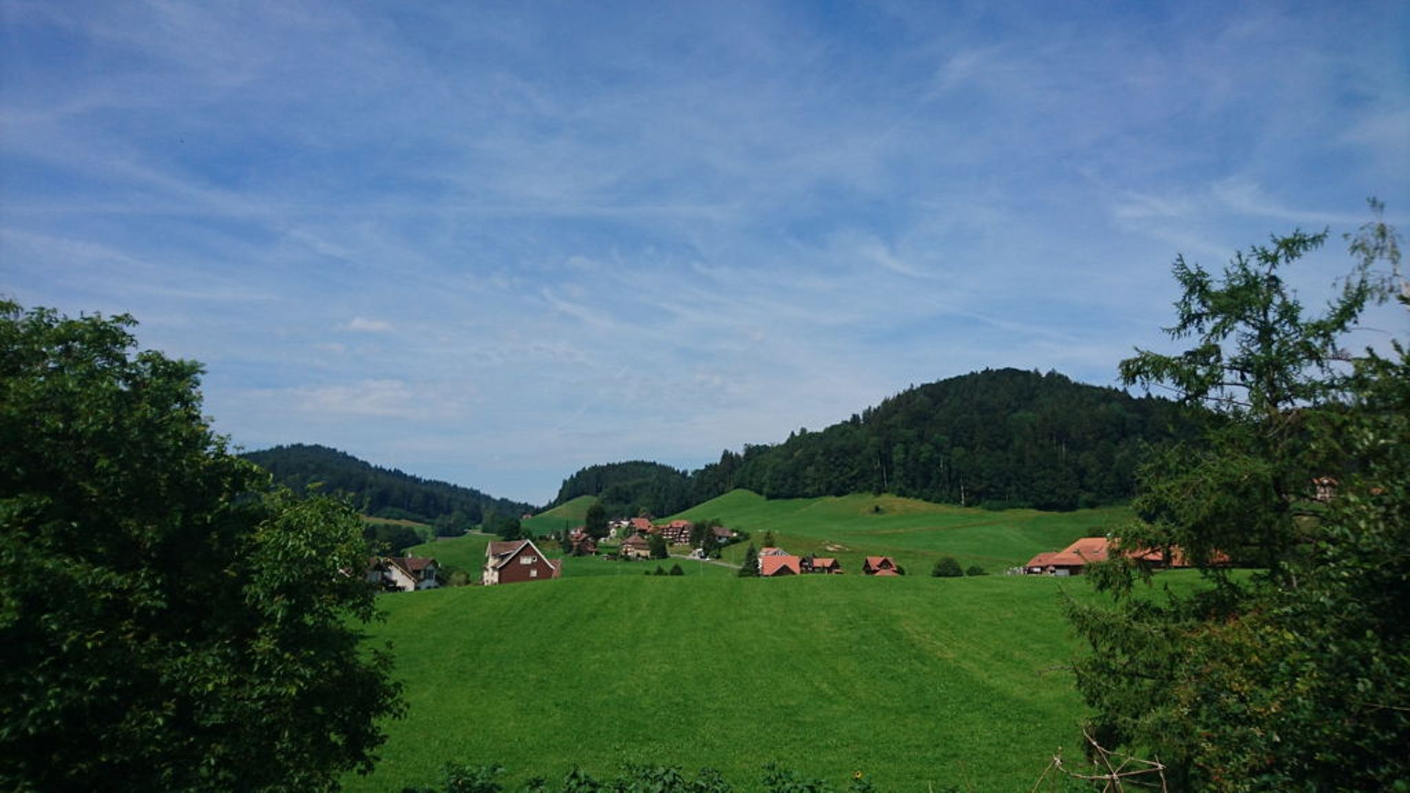 Paisagem idílica de aldeia com prados verdes e montanhas sob um céu azul.