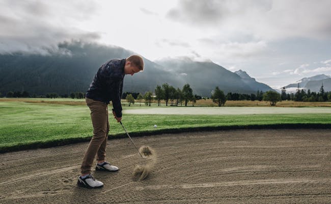 Golf Samedan, spelare slår från sandbunker, grönt område, berg i bakgrunden.
