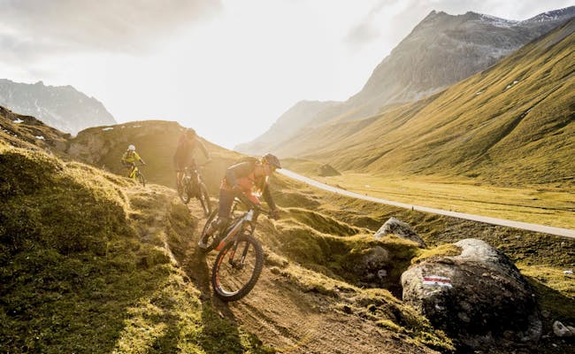Ciclistas de montanha no Trilho Albula, com pastagens verdes e montanhas ao fundo