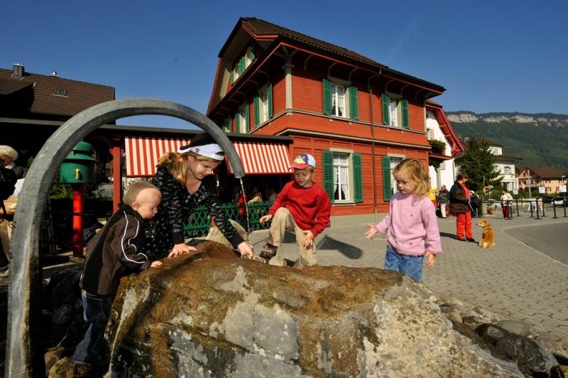 Les enfants jouent près de la cascade à Stans sous un ciel dégagé, avec en arrière-plan un bâtiment rouge.