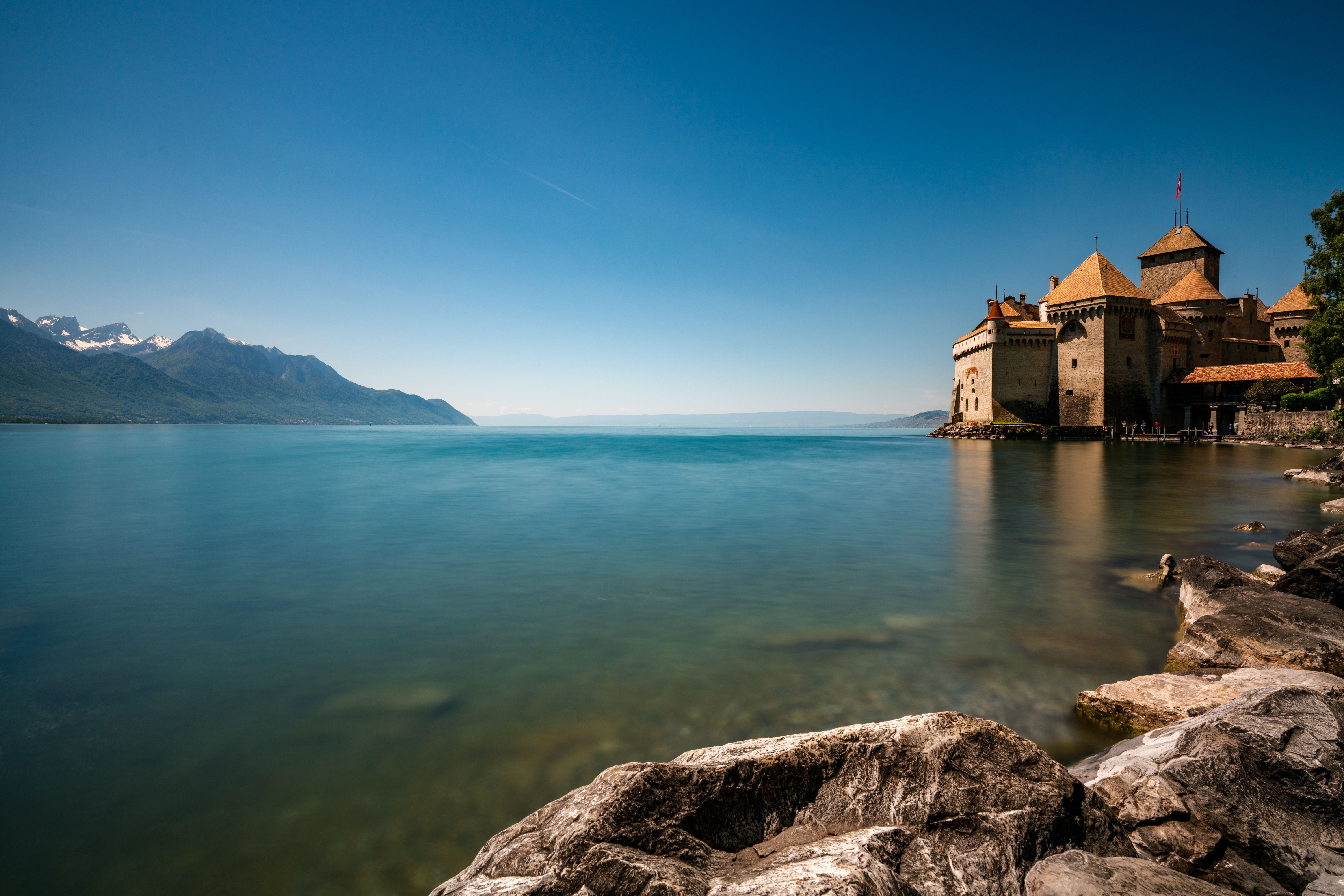 Chateau Chillon di Danau Geneva dengan permukaan air yang tenang dan Alps di latar belakang.