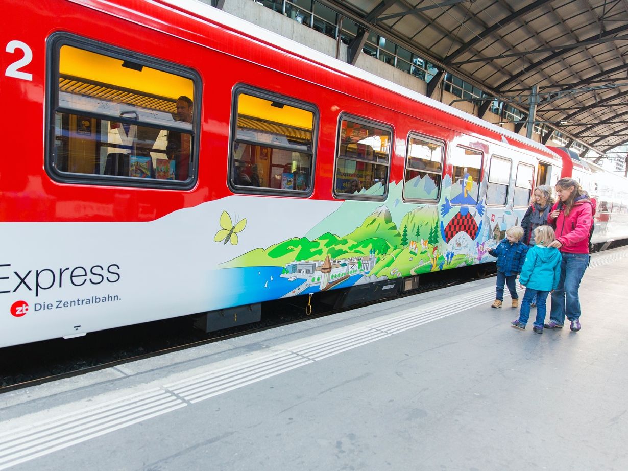 Keluarga dengan kereta di stesen, sedang menaiki kereta. Kereta berwarna merah dengan reka bentuk berwarna-warni.