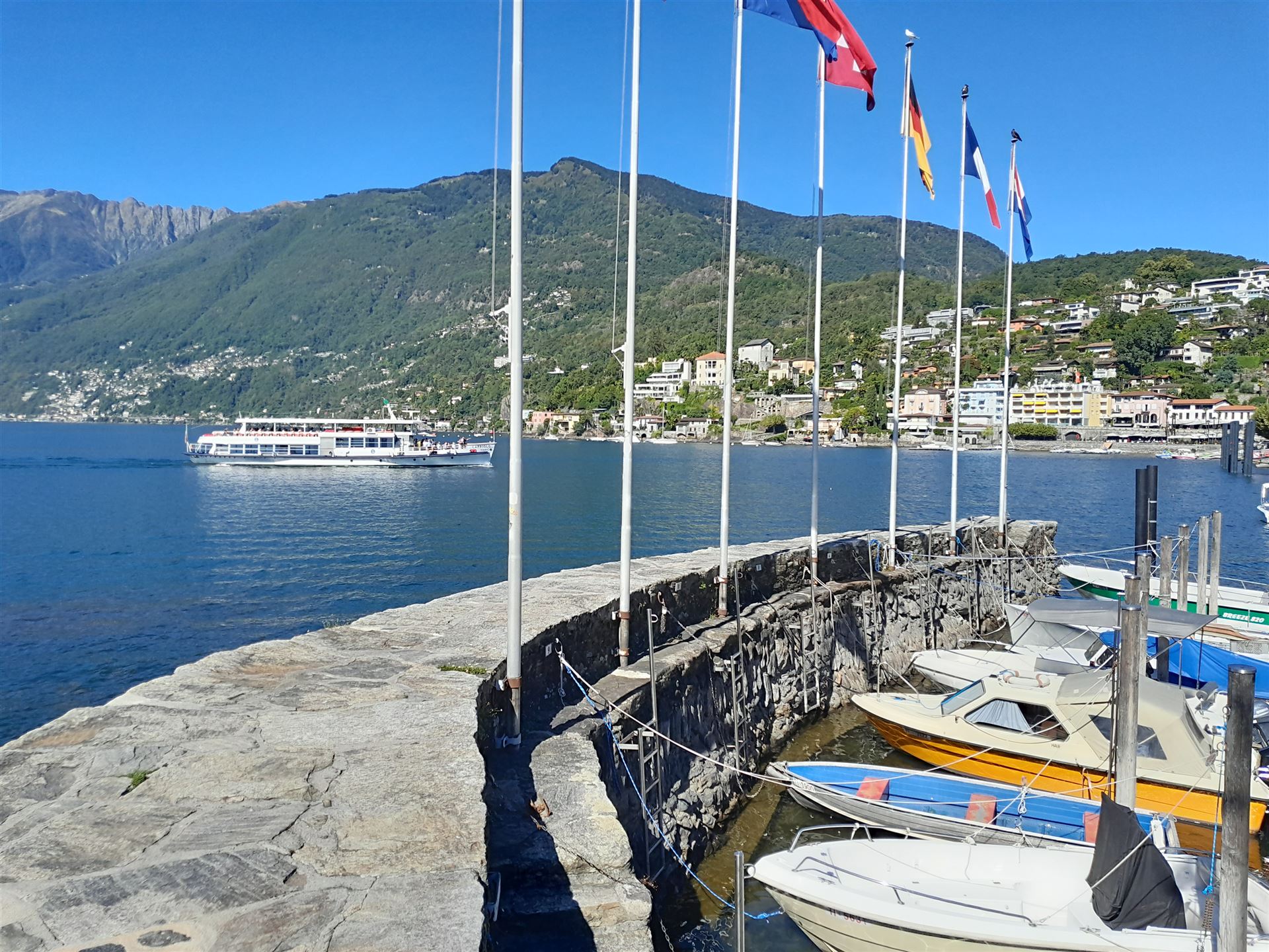 Ticinoquad: Vista del puerto en Ascona en el Lago Maggiore con barcos coloridos y montañas de fondo.