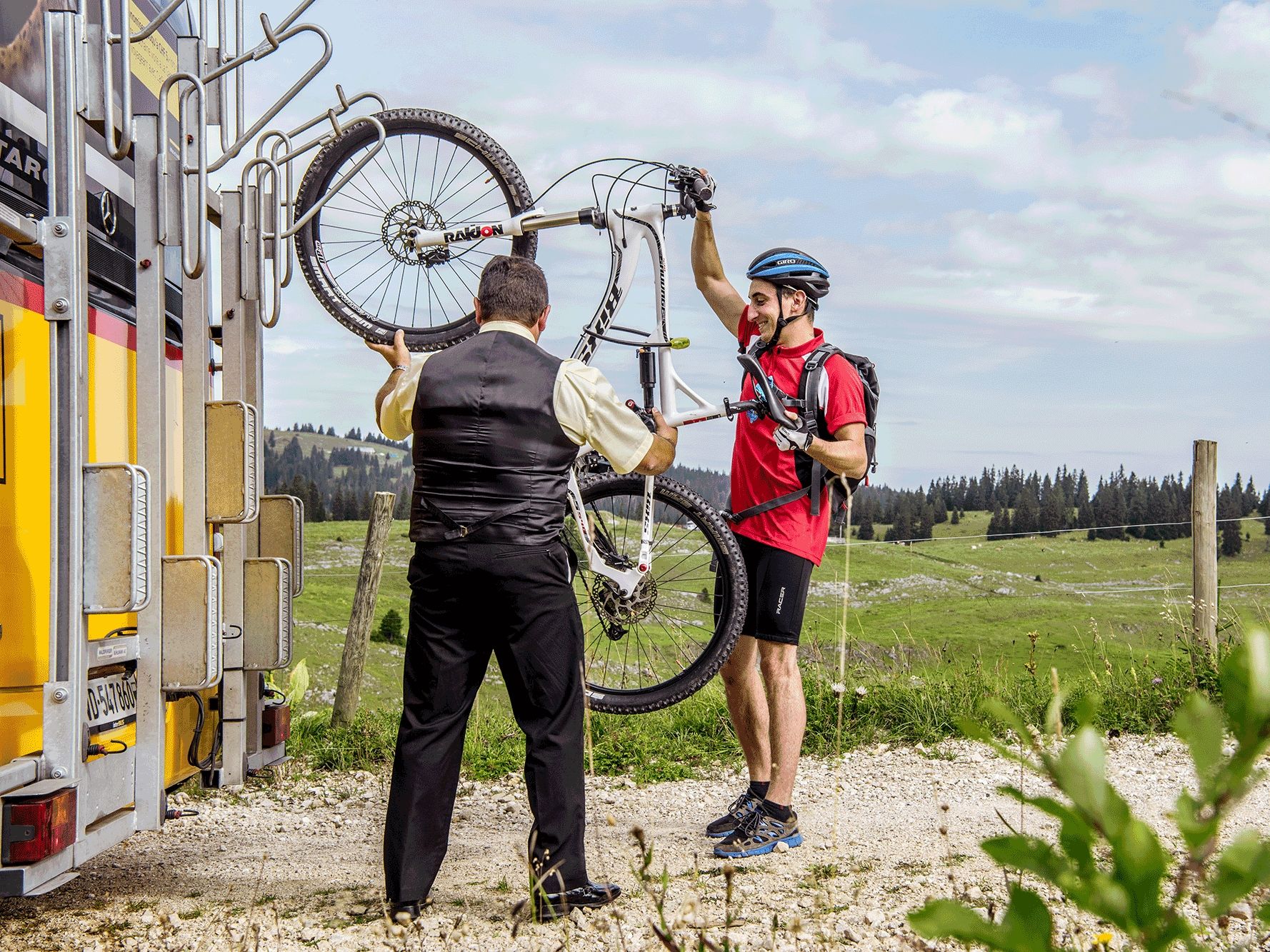การขนส่งจักรยานในสวิตเซอร์แลนด์: ชายคนหนึ่งยกจักรยานขึ้นไปใน Postauto โดยมีทุ่งหญ้าเขียวขจีและภูเขาเป็นฉากหลัง.