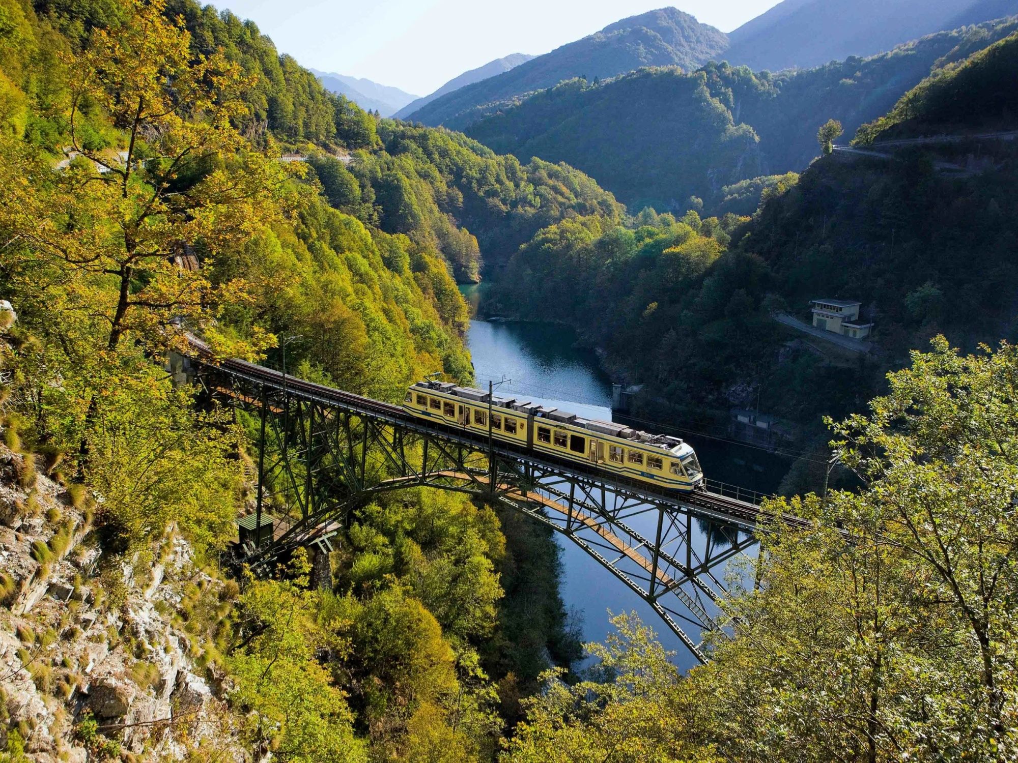 Centovalli Railway crosses a bridge in the green landscape. The surroundings are surrounded by trees.