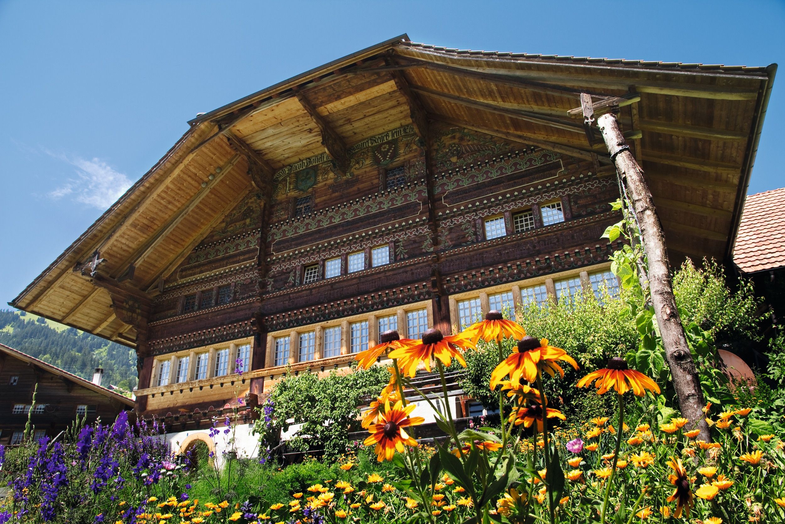 Housová cesta Simmentaler s tradičním dřevěným domem a pestrými květinami v popředí.