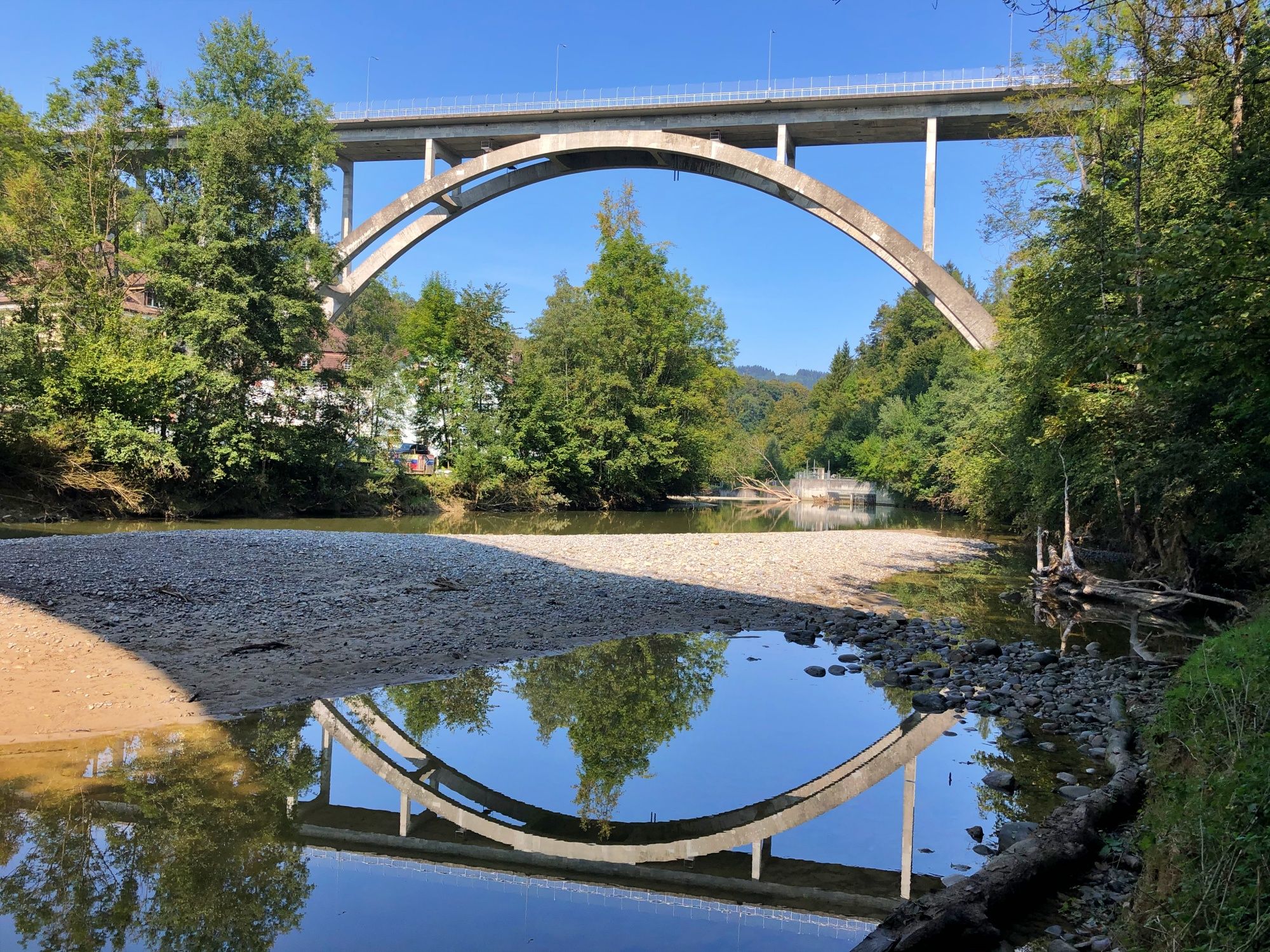 St. Galler Brückenweg ukazuje betonový most přes řeku, obklopený stromy a vodou. Za slunečného počasí.