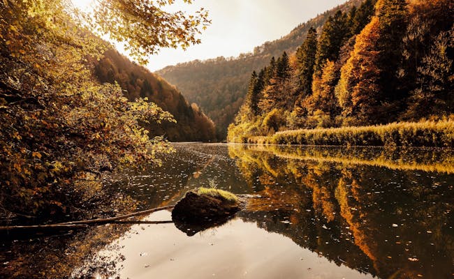 Parco Naturale di St Ursanne Doubs con foglie autunnali colorate lungo il fiume, la superficie tranquilla dell'acqua riflette il paesaggio.