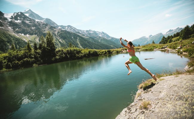 Saltando al agua en Blatten Schwarzsee, prados verdes, montañas al fondo, cielo claro.