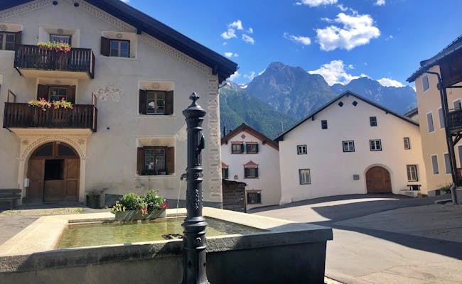 Piața principală din Sent cu fântâni, înconjurată de munți și clădiri istorice din Engadin.