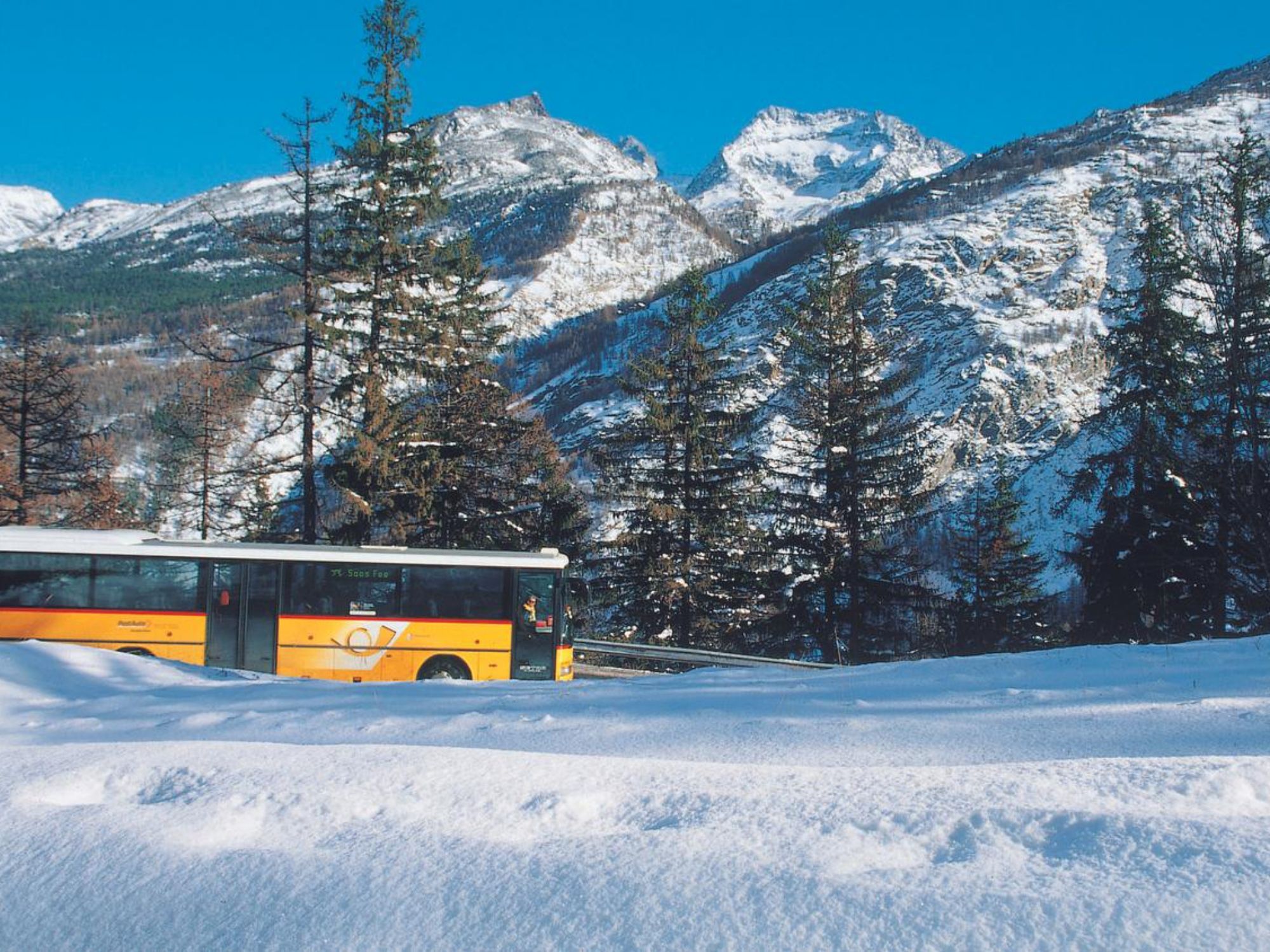 Σας Fee: Το Postauto χτυπάει το χιόνι, περιτριγυρισμένο από βουνά και κωνοφόρα δέντρα στο χειμερινό τοπίο.