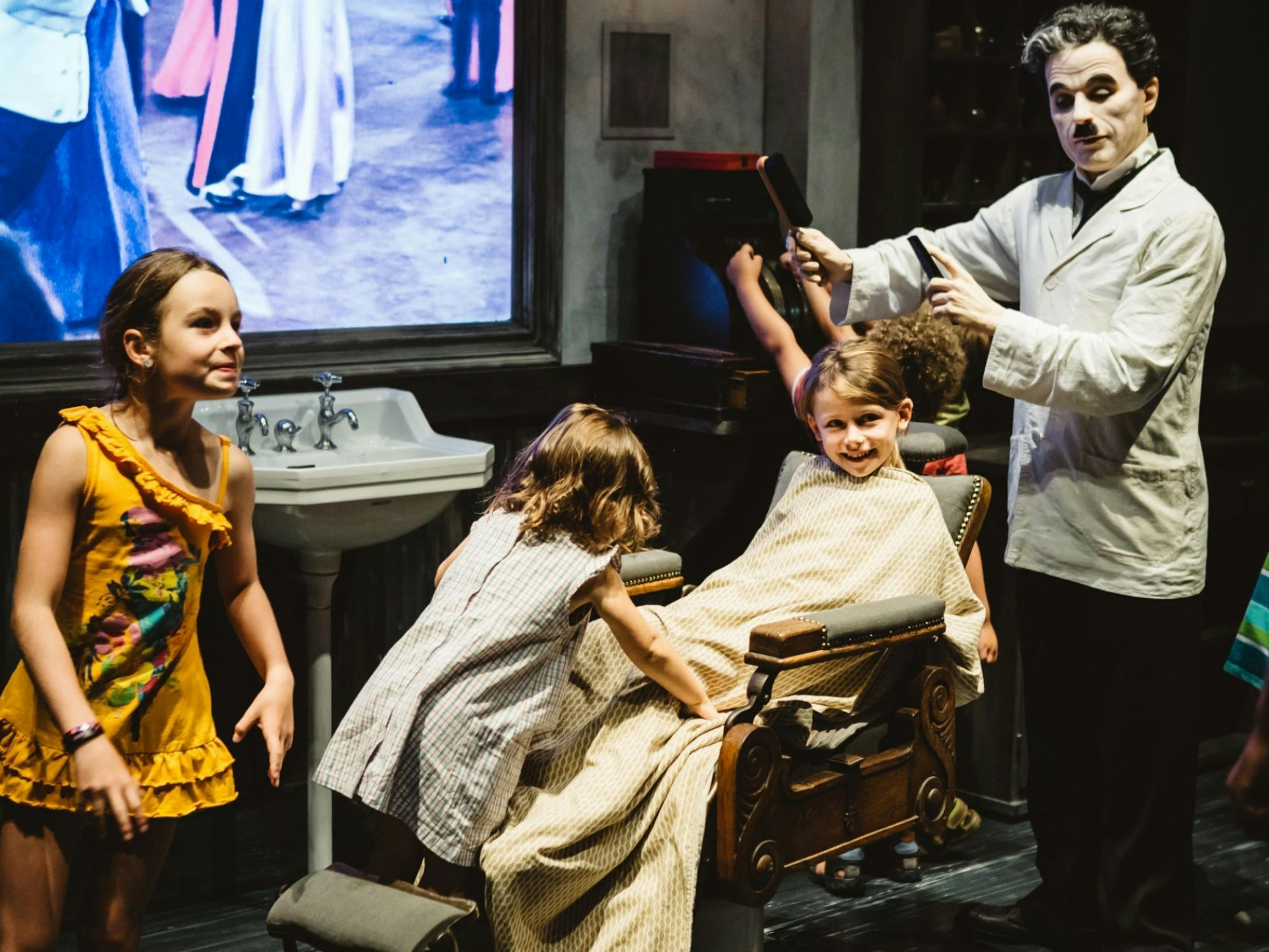 Chaplin als Barbier in einem Museum mit fröhlichen Kindern und historischem Ambiente.