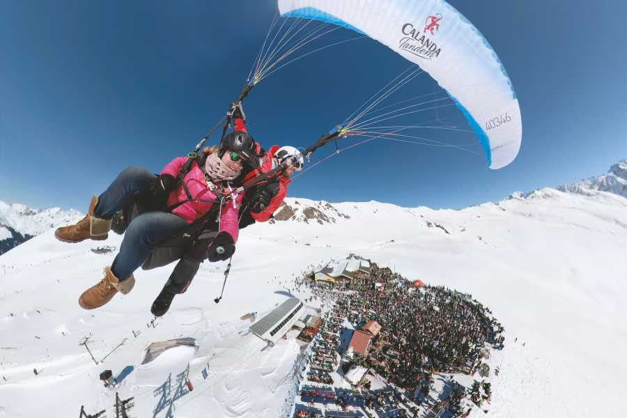Thermikflug in Davos beim Paragliding über schneebedeckte Berge und eine fröhliche Menschenmenge