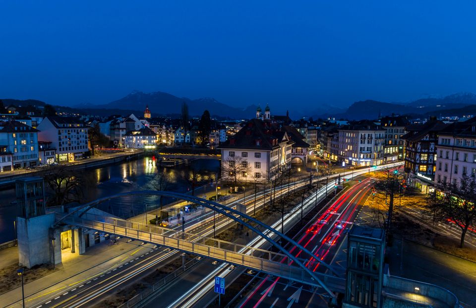 Luzern bei Nacht mit beleuchteten Gebäuden, Straßenverkehr und Bergen im Hintergrund
