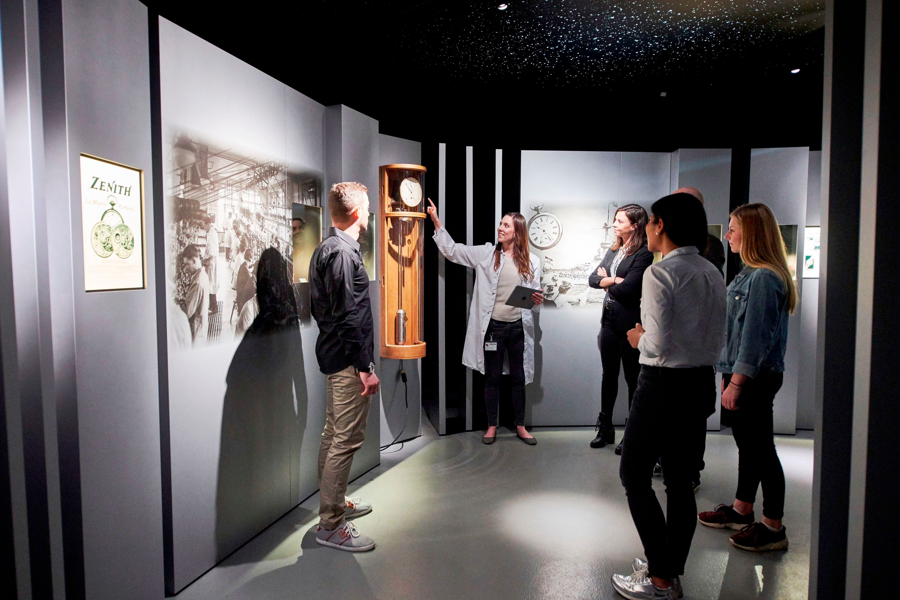 Zenith-Ausstellung - Besucher hören Informationen zur Uhrentechnik. Sichtbar sind Uhren und historische Fotos.