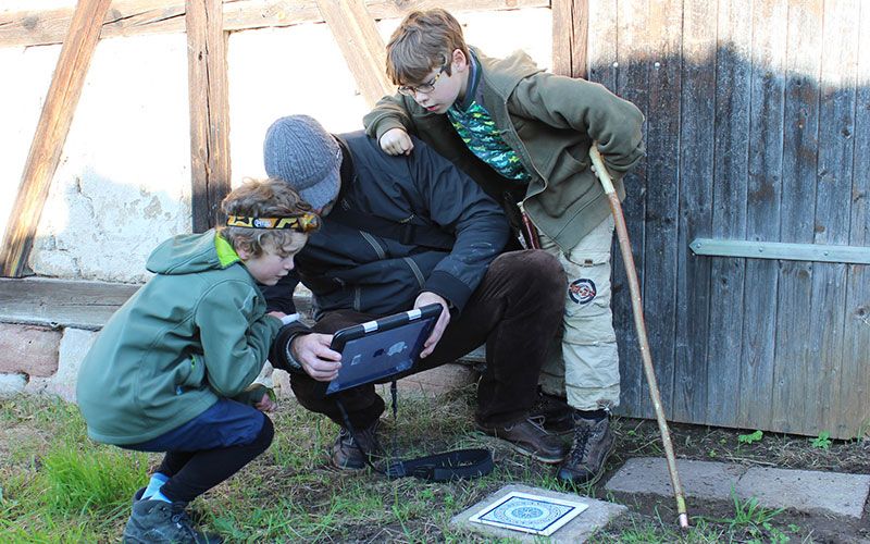 Familie spielt Escape Game im Freien, mit Tablet und Rätseln.