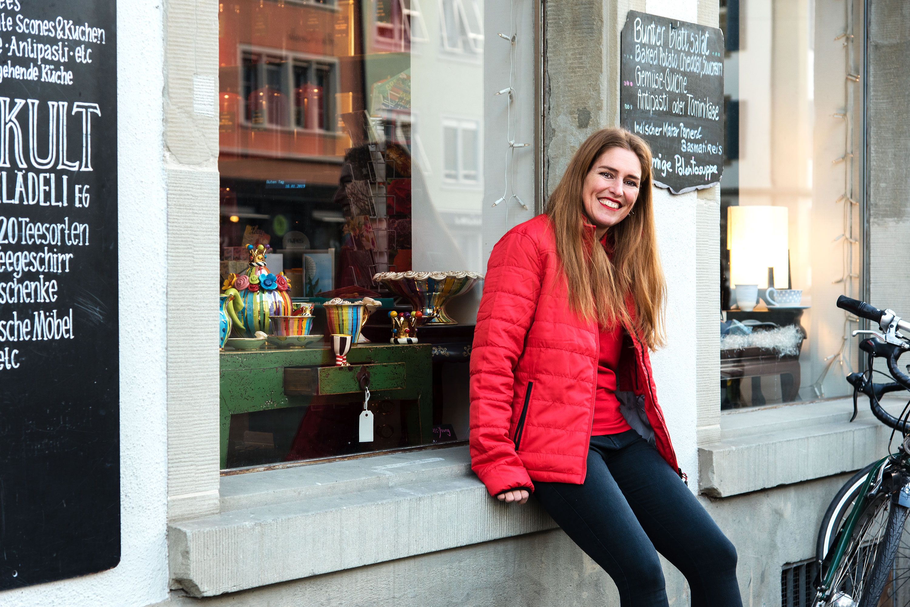 Seorang wanita memakai jaket merah duduk di hadapan jendela kedai yang dihiasi dengan barang hiasan berwarna-warni.