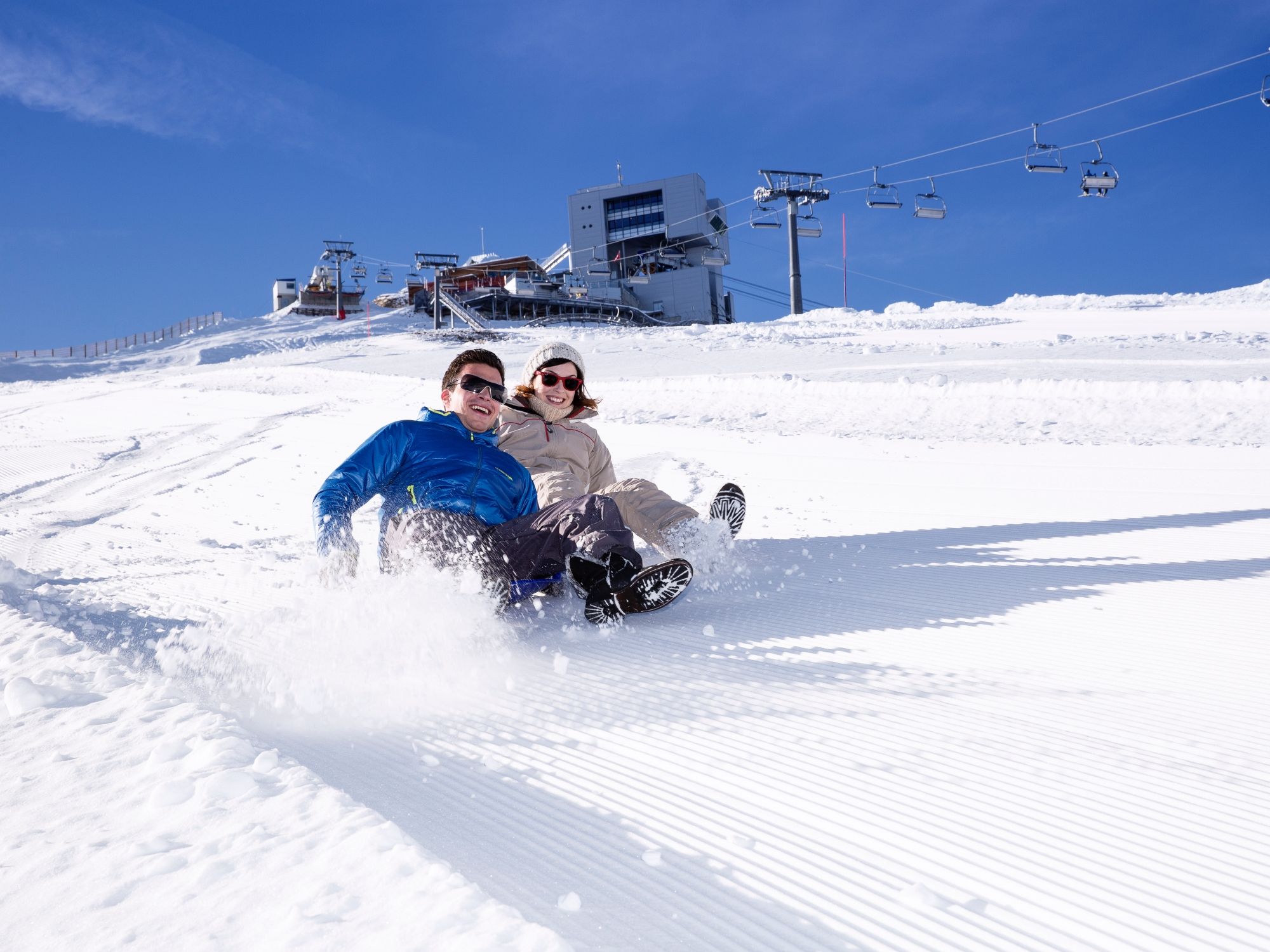 펀 파크 슬리첼: 두 사람이 눈 위에서 썰매를 타고 있고, 배경에는 의자 리프트가 있습니다.