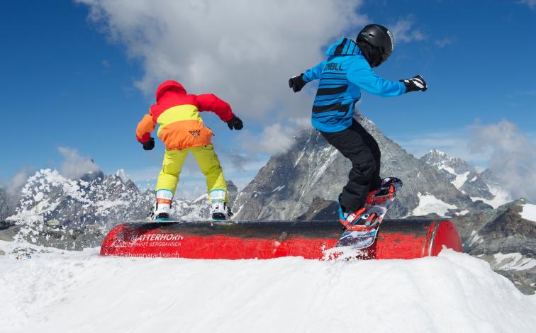 การเรียนสโนว์บอร์ดส่วนตัวสำหรับเด็กในเซอร์แมตต์แสดงให้เห็นเด็กสองคนบนสโนว์บอร์ด เด็กๆ สวมใส่เสื้อผ้าสีสันสดใสและยืนอยู่บนกระโดด มีภูเขาที่มองเห็นได้และท้องฟ้าสีฟ้าอยู่เบื้องหลัง