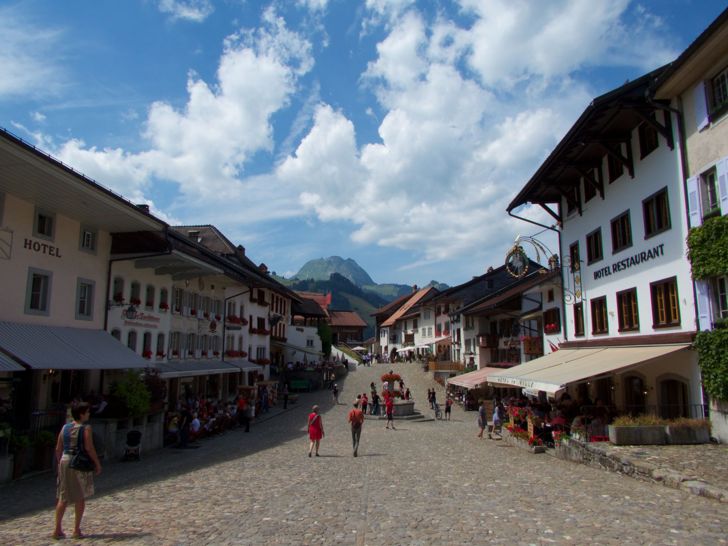 Gruyeres: Një rrugë idilike me ndërtesa, turistë dhe male në sfond, kënaqësi verore.