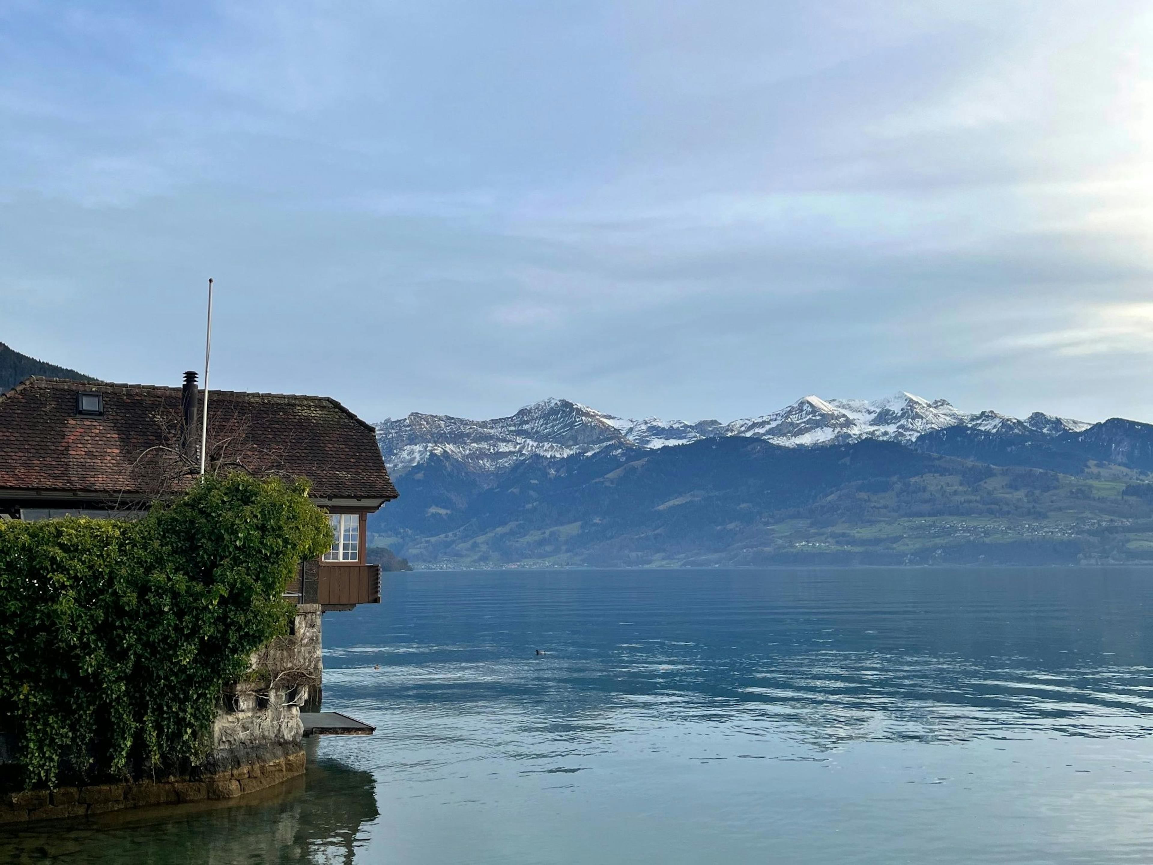 Gunten: Blick auf den See mit einem malerischen Gebäude und schneebedeckten Bergen im Hintergrund.