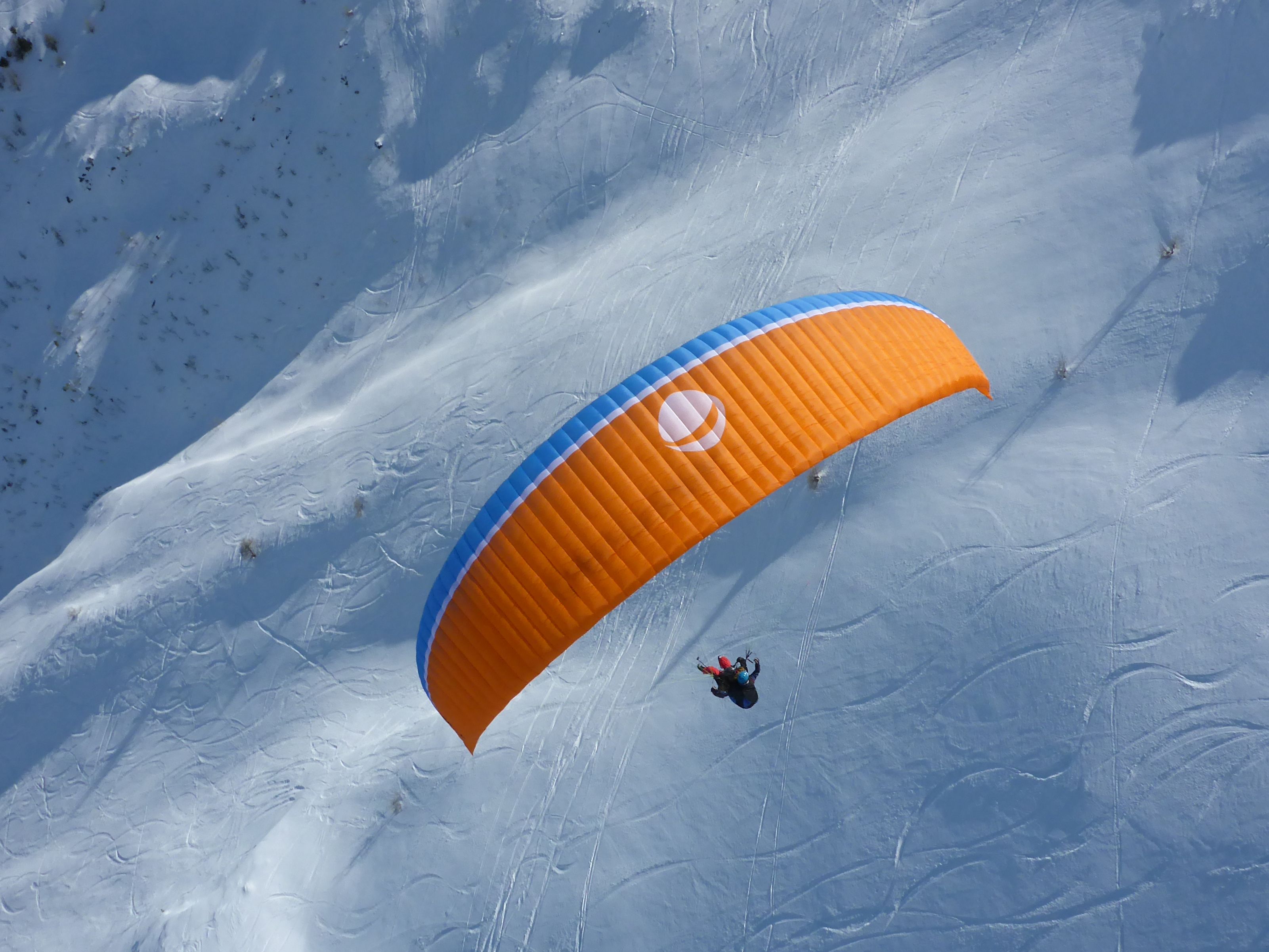 Paragliding nad zasněženou krajinou v Davosu v zimě, lyžařské středisko, aktivní osoba s oranžovo-modrým křídlem