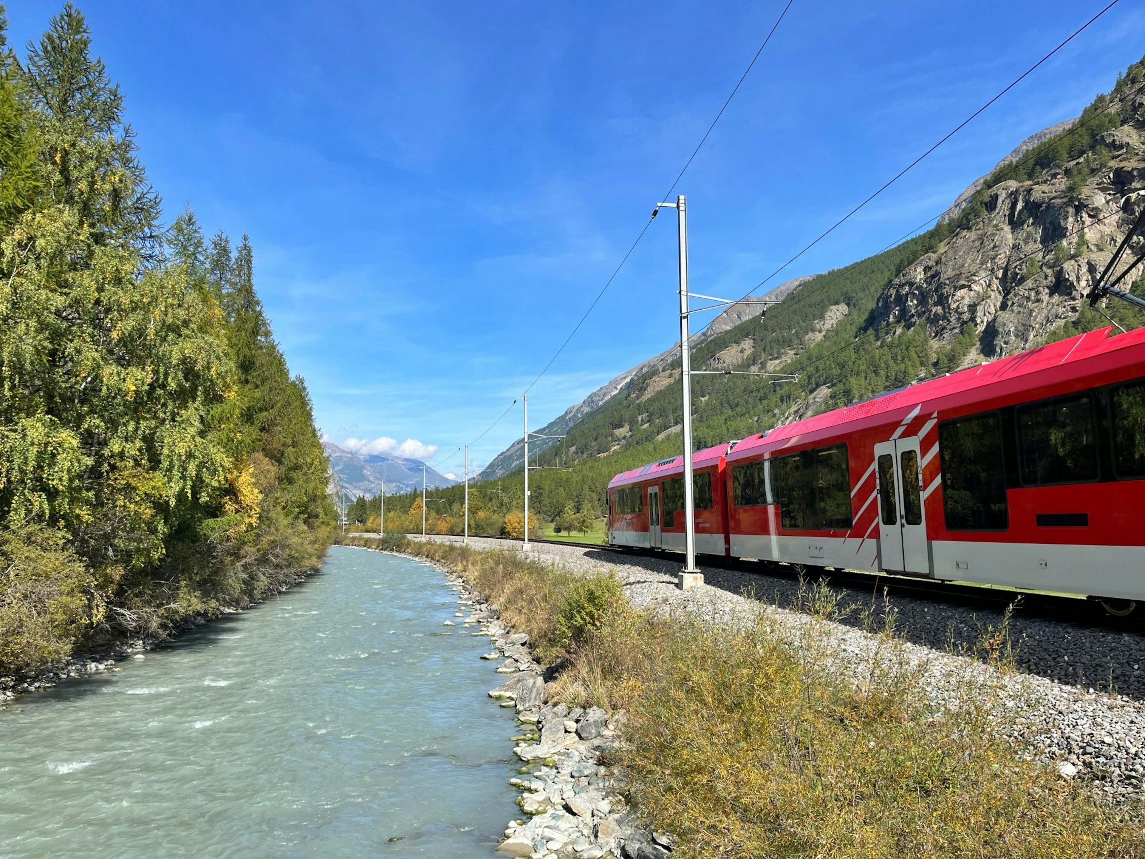Treno: Treno rosso lungo il fiume con montagne sullo sfondo, ideale per gli amanti della natura in estate.