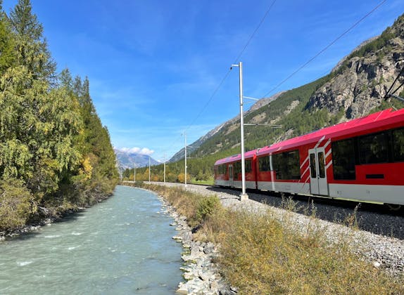 Treno: Treno rosso lungo il fiume con montagne sullo sfondo, ideale per gli amanti della natura in estate.