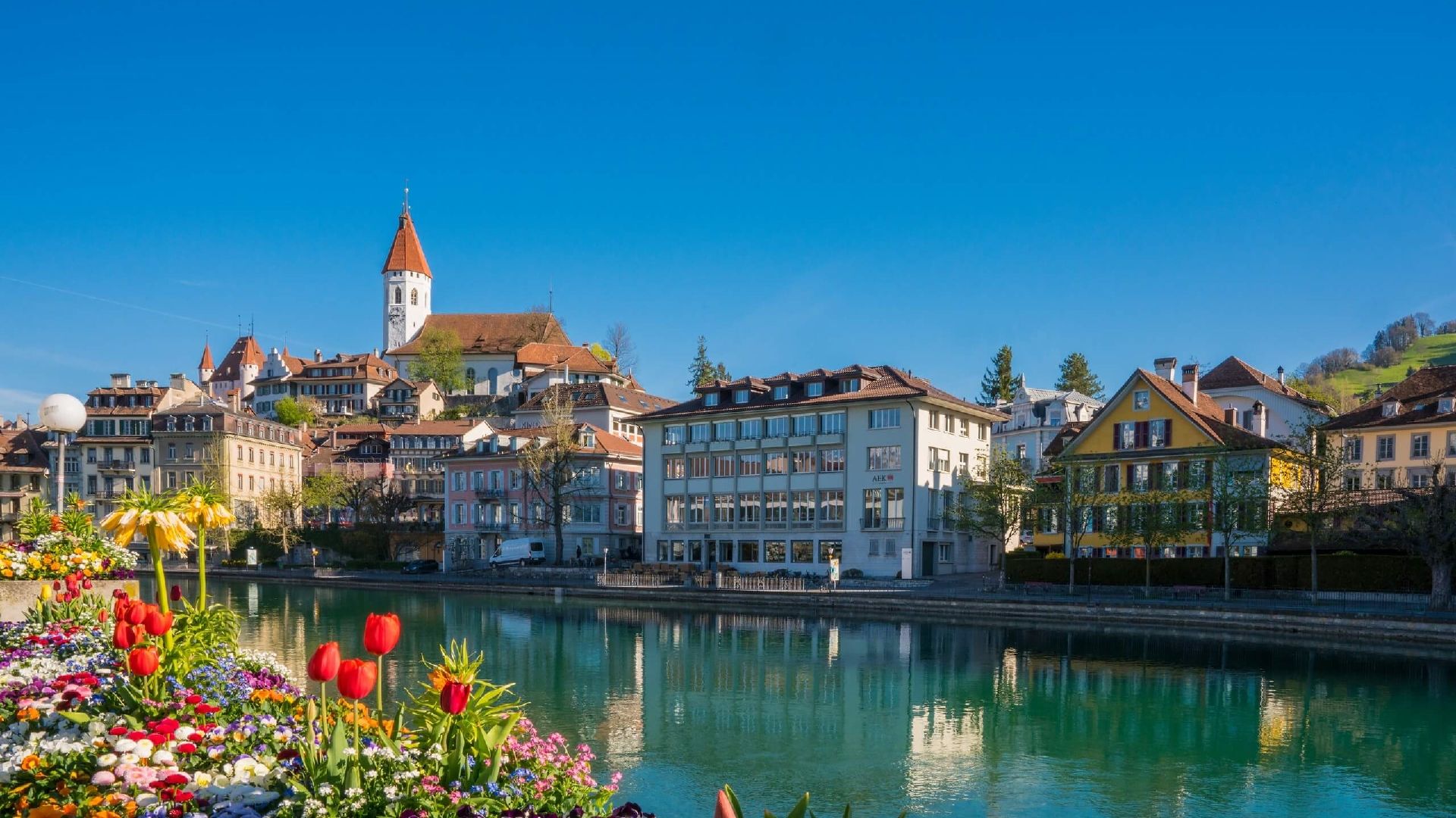 Pandangan ke Thun dengan bunga berwarna-warni di sepanjang tepi tasik, langit cerah, seni bina bersejarah.