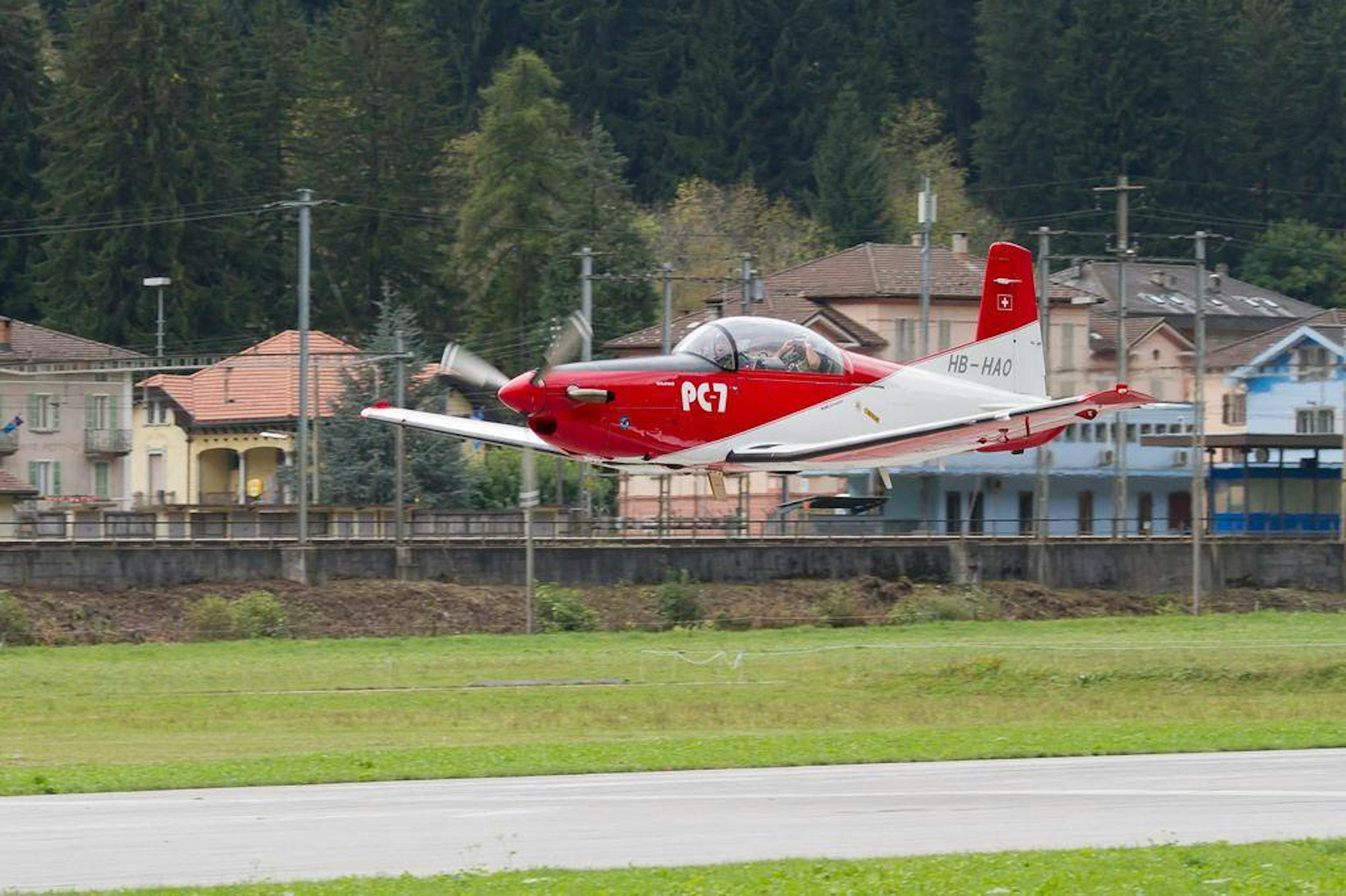 Pilatus PC-7 takes off over a meadow, surrounded by trees and houses.