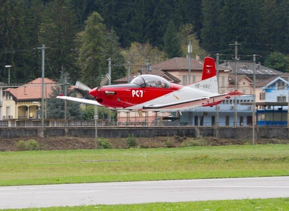 Pilatus PC-7 vzlet z louky obklopené stromy a domy.