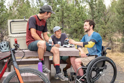 Rollstuhl: Gruppe von Freunden beim Picknick in der Natur mit Mountainbikes und Getränken.