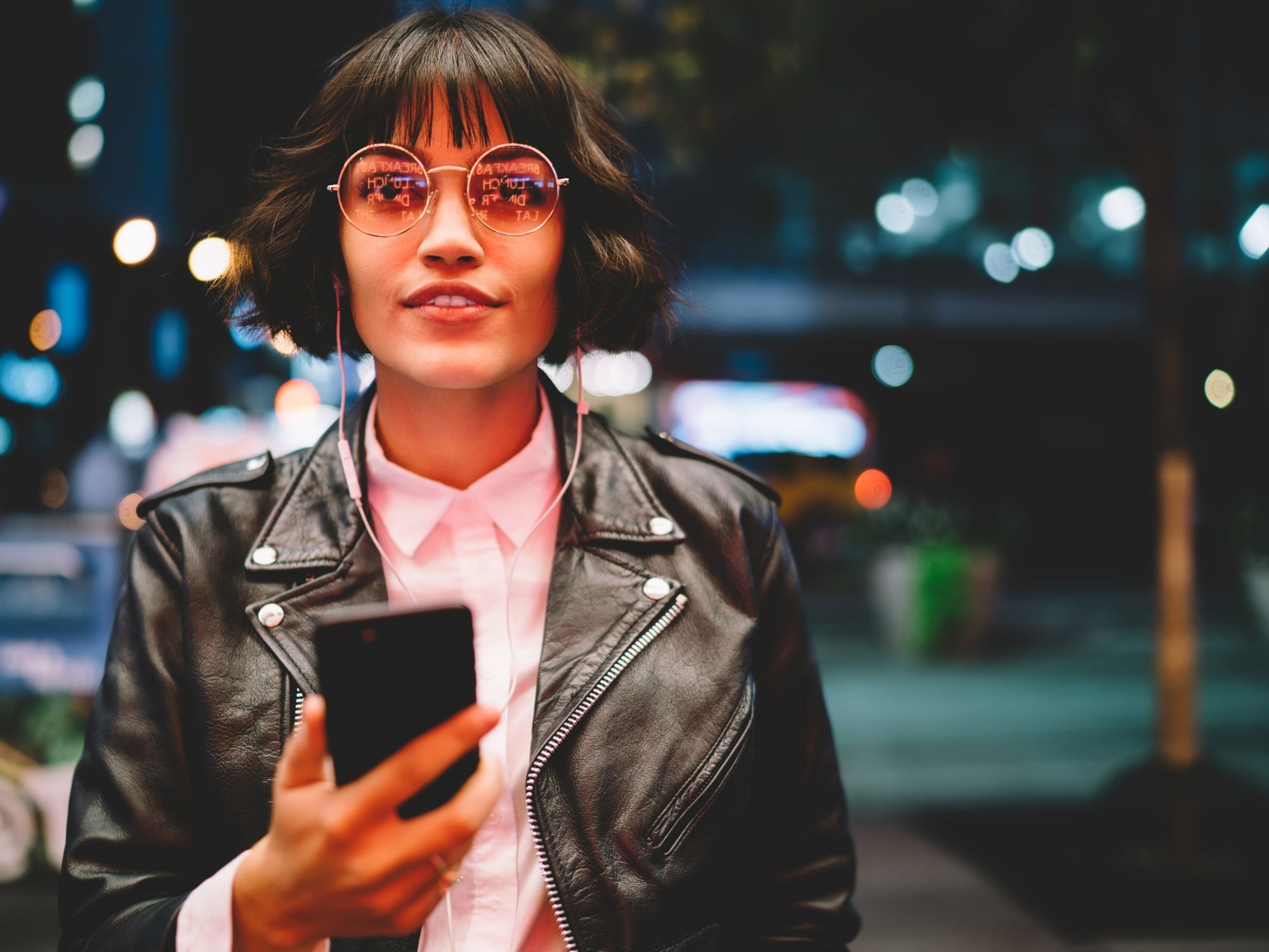 Sato Code: Junge Frau mit Handy in glamourösem Outfit in belebter Umgebung bei Nacht.