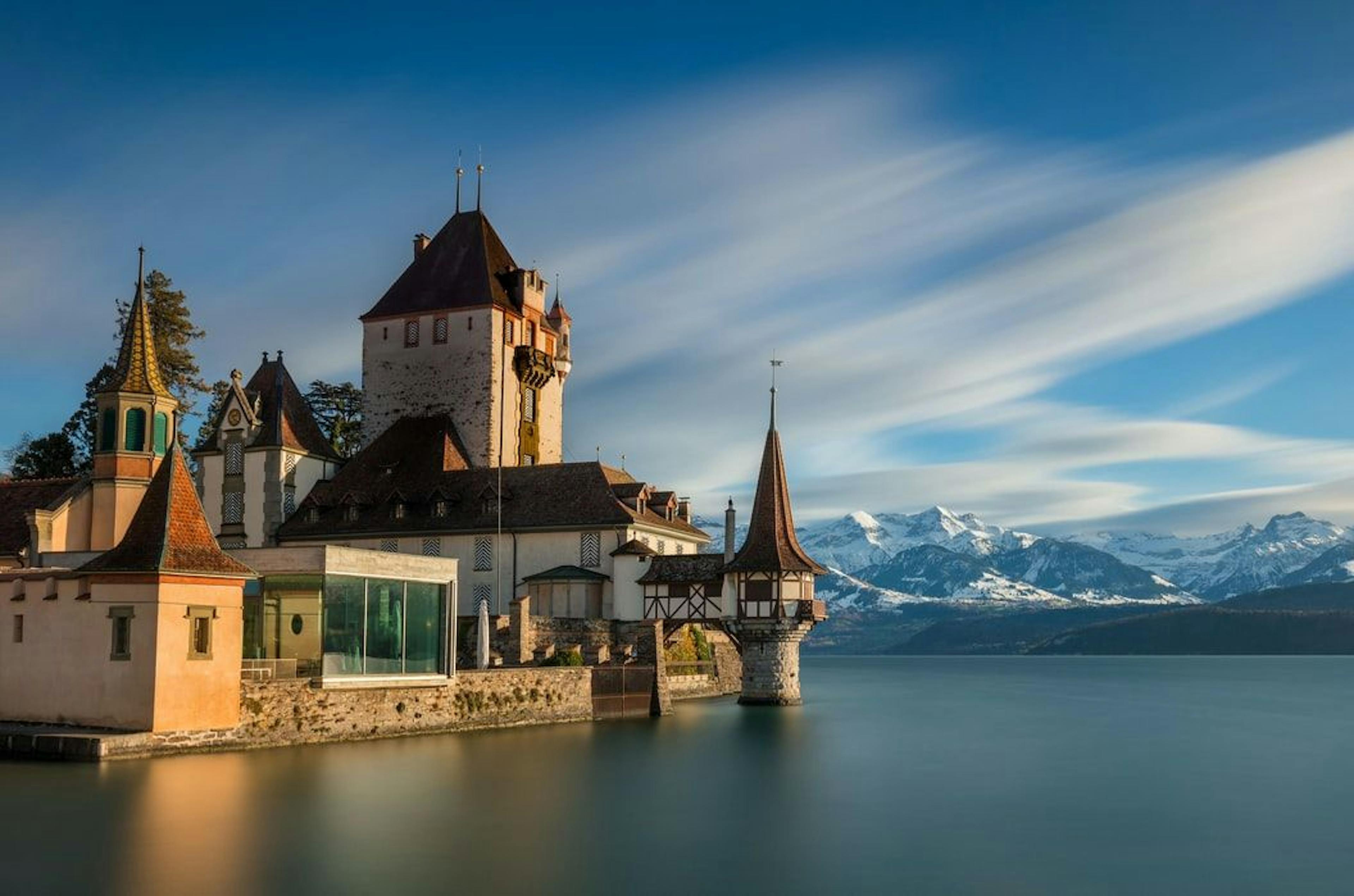 Istana Oberhofen di Danau Thuner dengan gunung bersalju di latar belakang.
