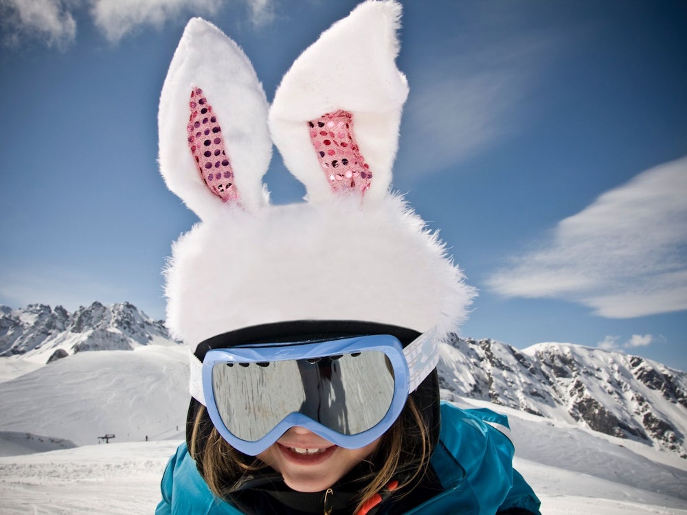 Заяц в снегу: ребёнок в лыжных очках и заячьих ушах зимой на Пизоле перед заснеженными горами.