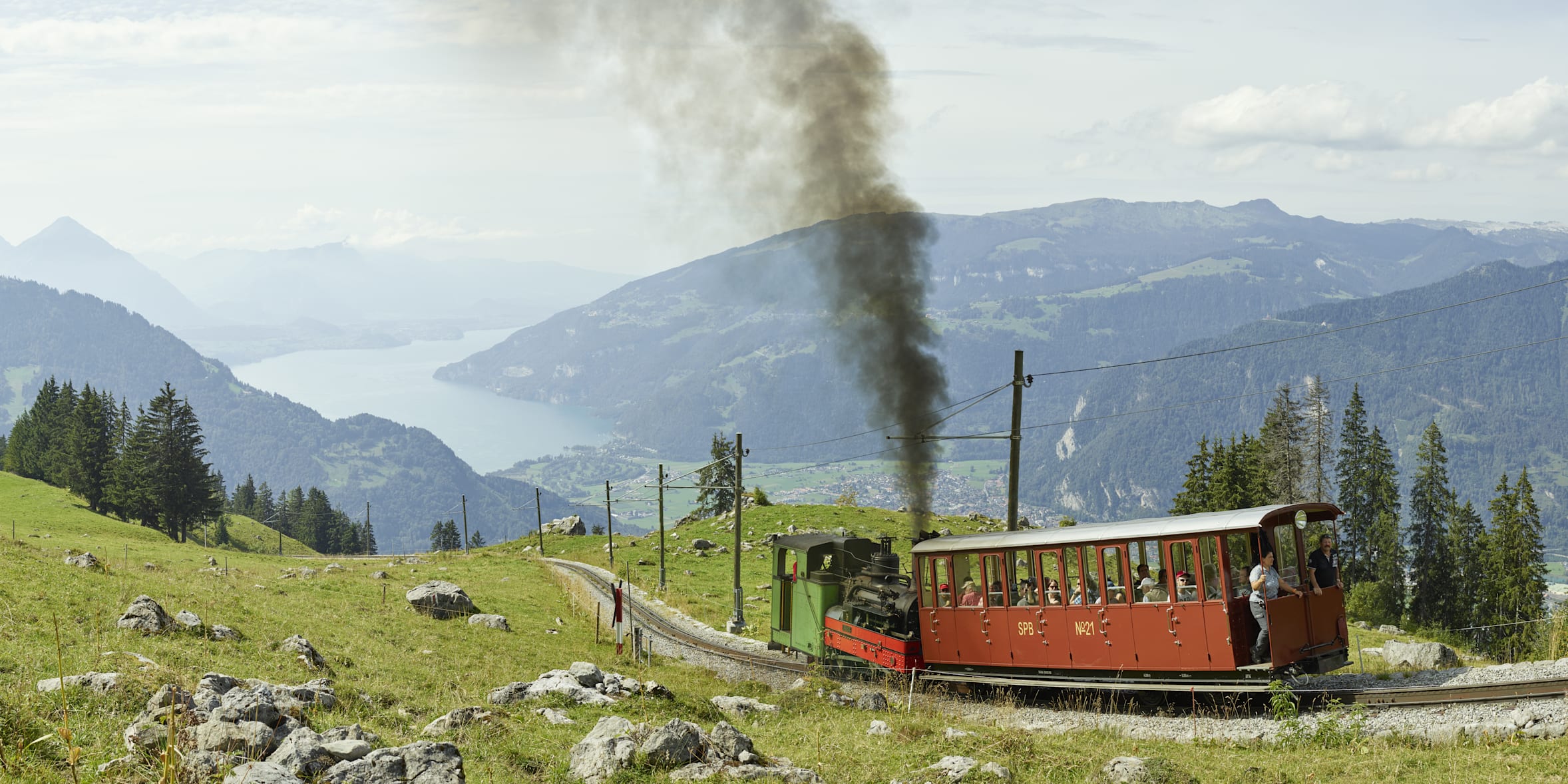 Schynige Platte Zahnradbahn Dampflokomotive dampft durch grüne Wiesen mit See im Hintergrund.