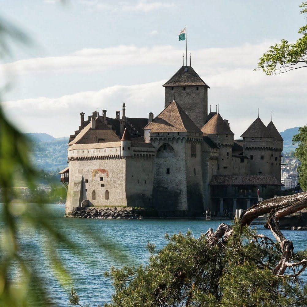 Chateau Chillon di Danau Geneva dengan seni bina zaman pertengahan dan pokok hijau di latar depan.
