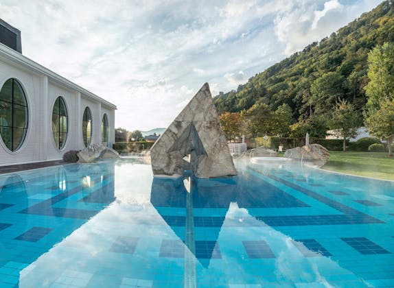 Θερμαλισμός Ταμίνα με επιφάνεια νερού και βράχους στο παρασκήνιο, περικυκλωμένο από πράσινο τοπίο.