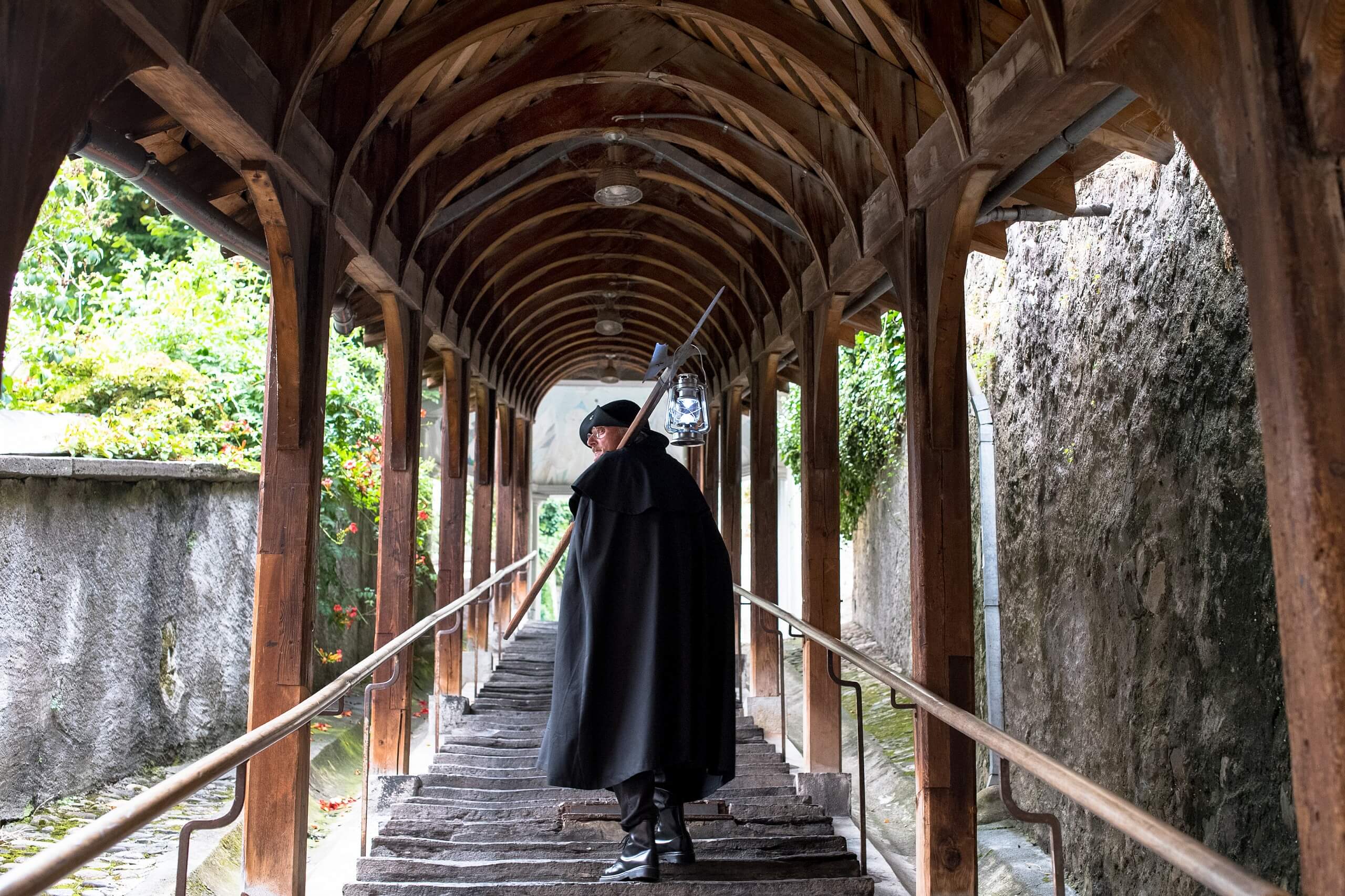 Stadswandeling Griezelig Thun met een persoon in een donkere mantel, die een lantaarntje vasthoudt en langs een houten brug loopt.