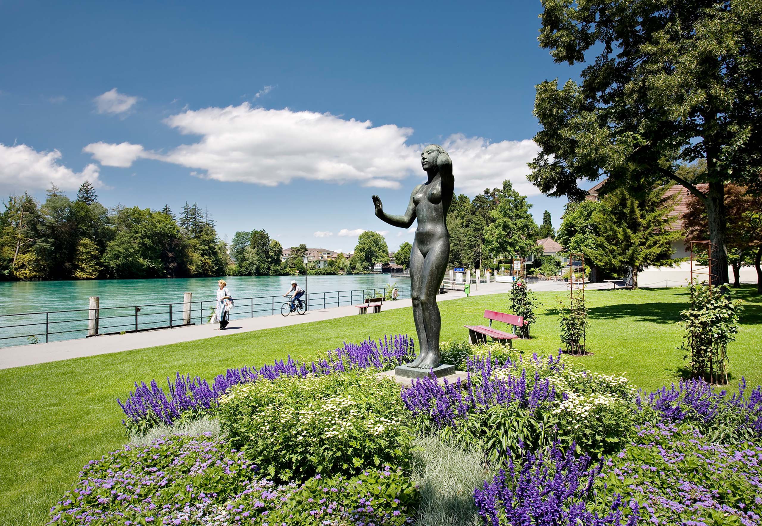 Thun Brahms Statue im Park am Thunersee mit Blumen und Radfahrern im Hintergrund bei schönem Wetter.